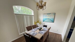 A dining area with a wooden table, chairs, a chandelier, a window with blinds, and a painting on the wall. - Lakeview Villa 5 Bedroom Home (Davenport)