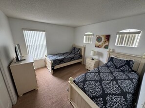 Een slaapkamer met twee eenpersoonsbedden, een nachtkastje met lamp, een dressoir en een raam met jaloezieën.