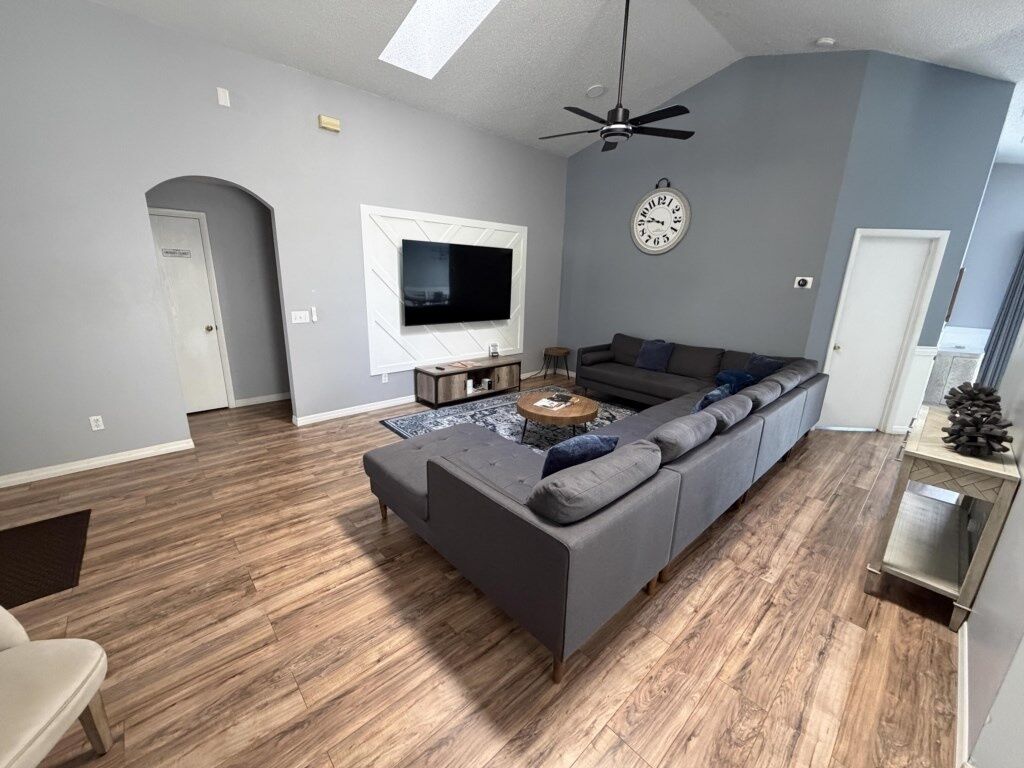 Un salon moderne avec un canapé d’angle, un téléviseur à écran plat, un ventilateur de plafond et une horloge murale.