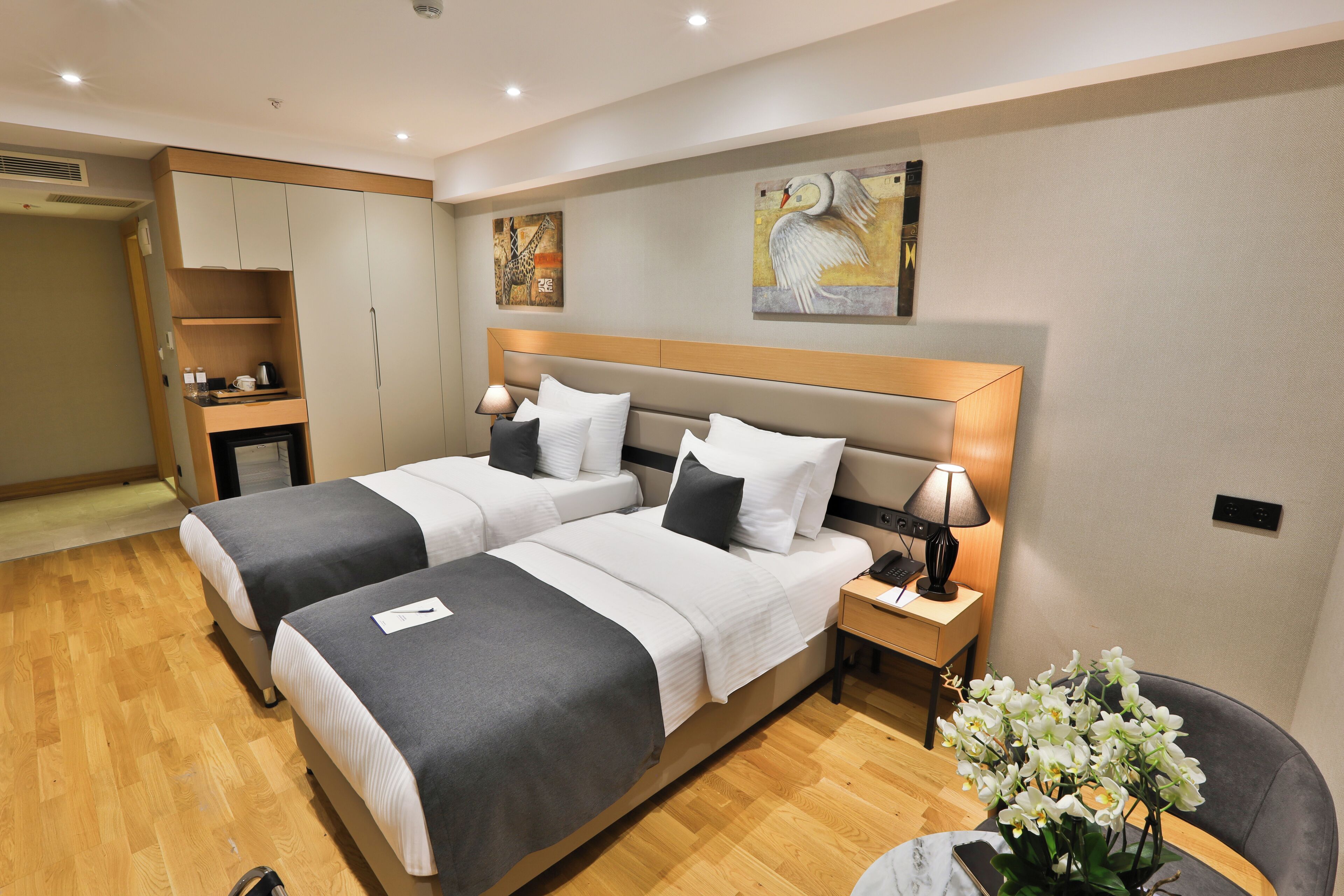 A hotel room with two beds, a nightstand with a lamp, a chair, and a small table with a vase of flowers.