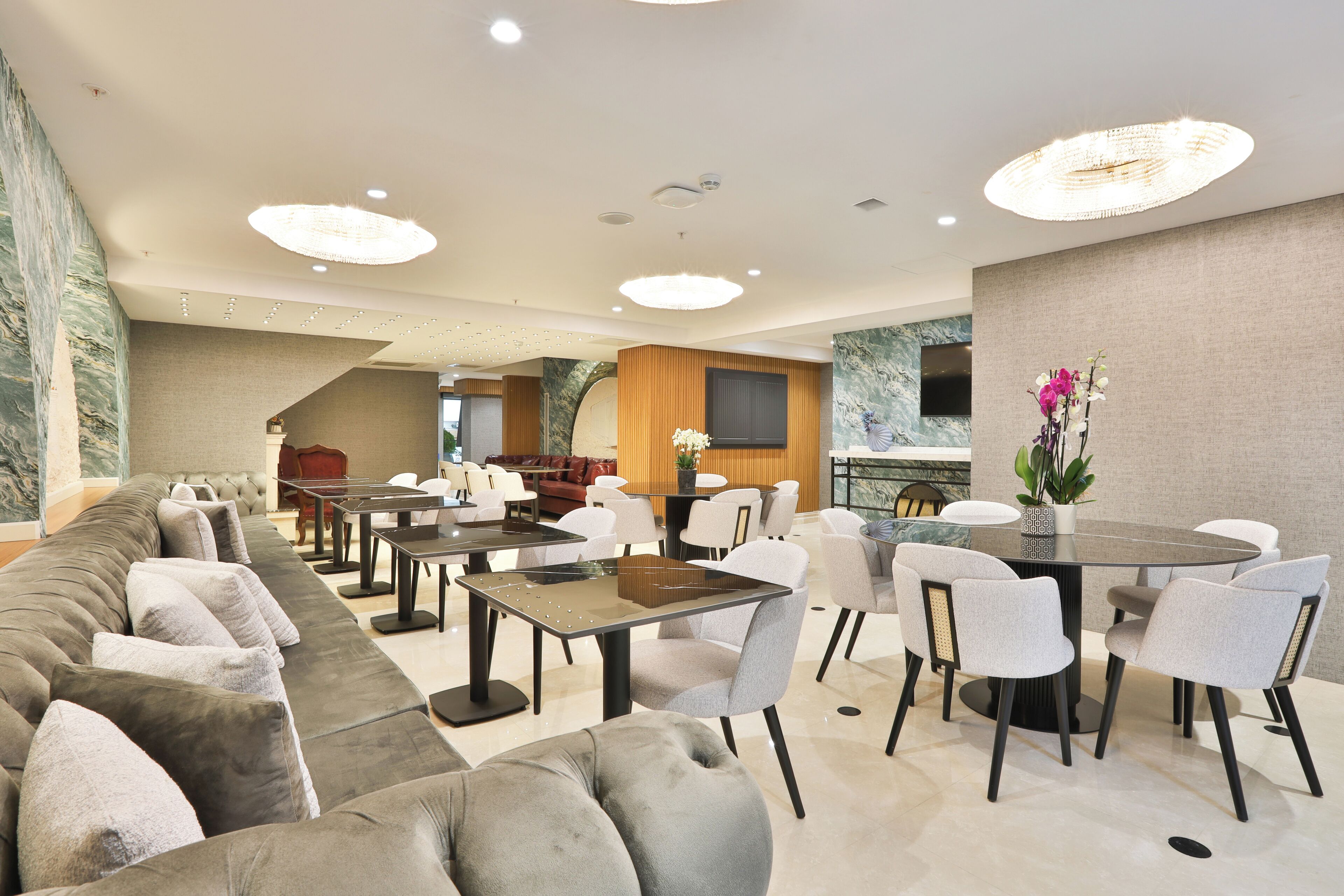 A modern hotel lobby with a long grey sofa, a round dining table with chairs, and a wooden accent wall.