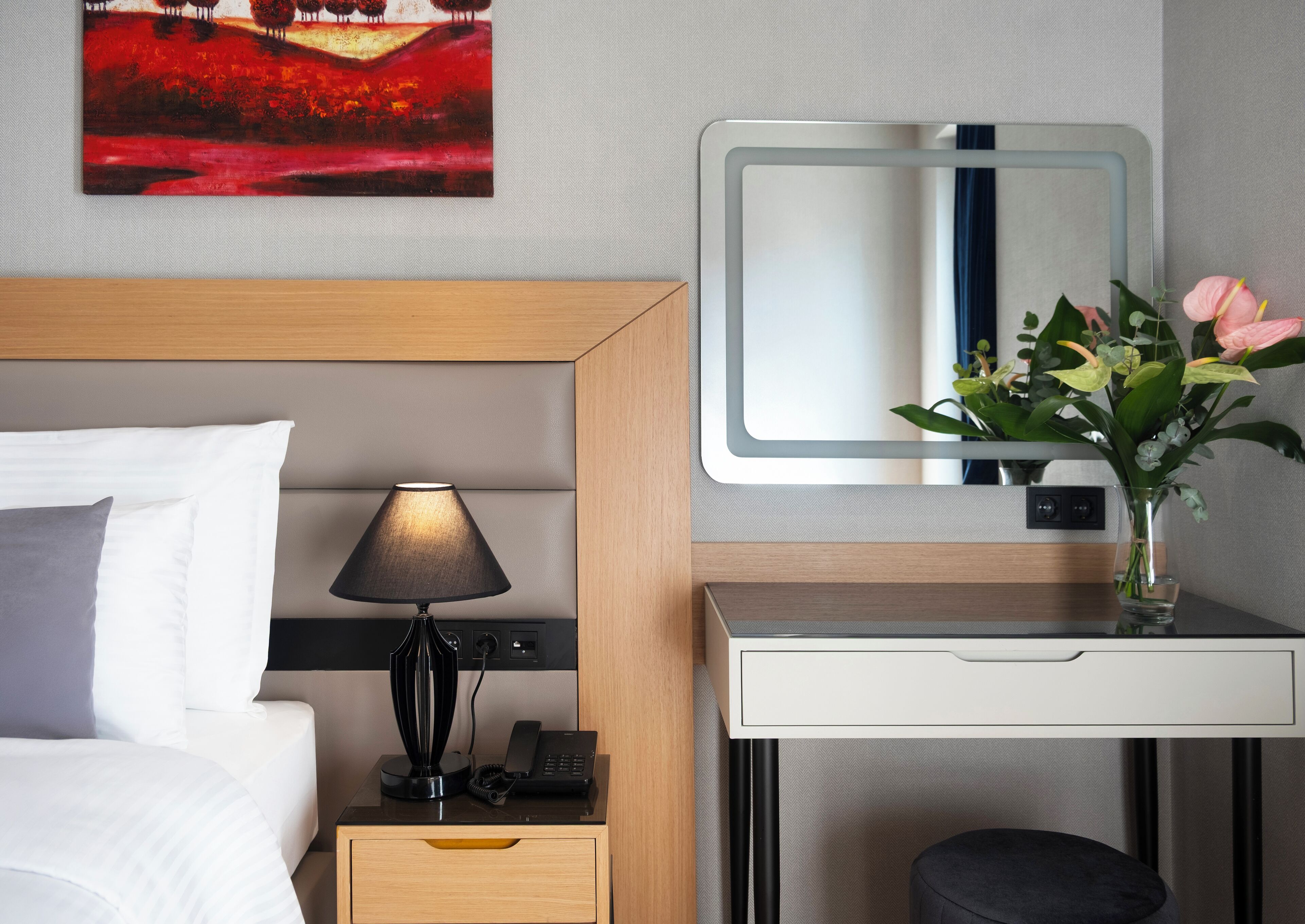 Una habitación de hotel con cama, lámpara de noche, escritorio, espejo y un cuadro en la pared.