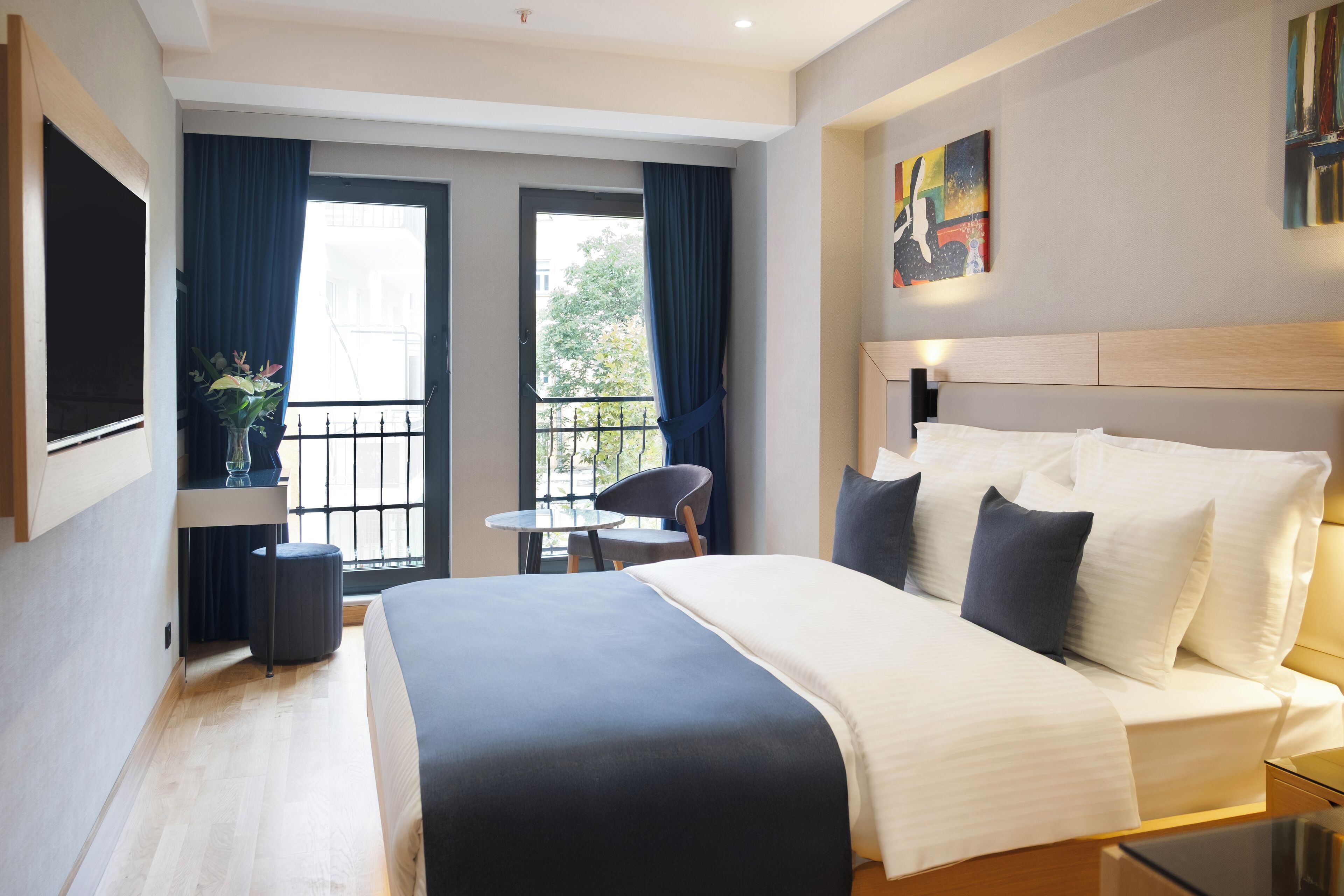A modern hotel room with a large bed, a small table, a chair, and a balcony with curtains.
