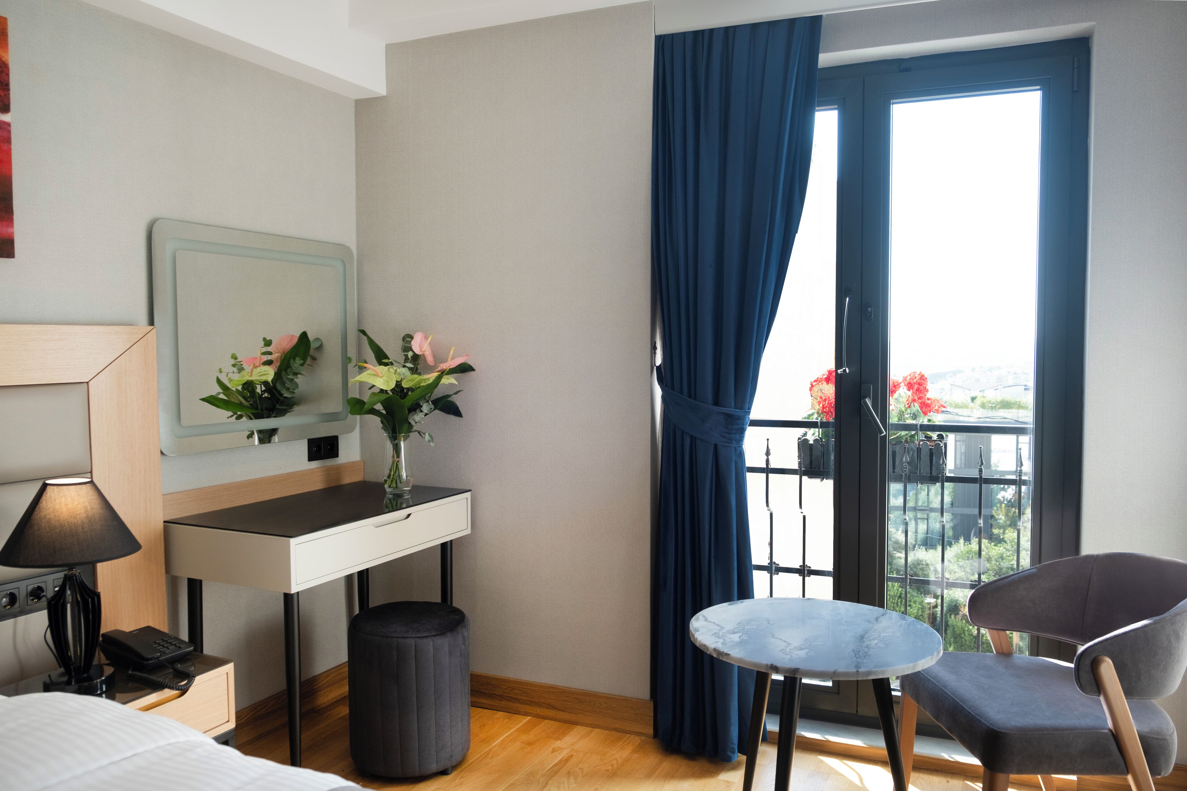 A hotel room with a bed, a desk with a lamp, a small table, a chair, a window with curtains, and a view of greenery outside.