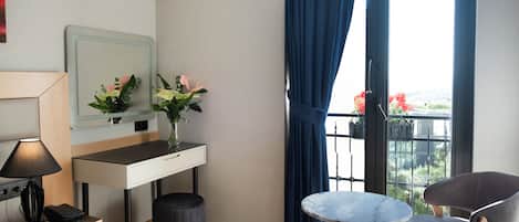 A hotel room with a bed, a desk with a lamp, a small table, a chair, a window with curtains, and a view of greenery outside.