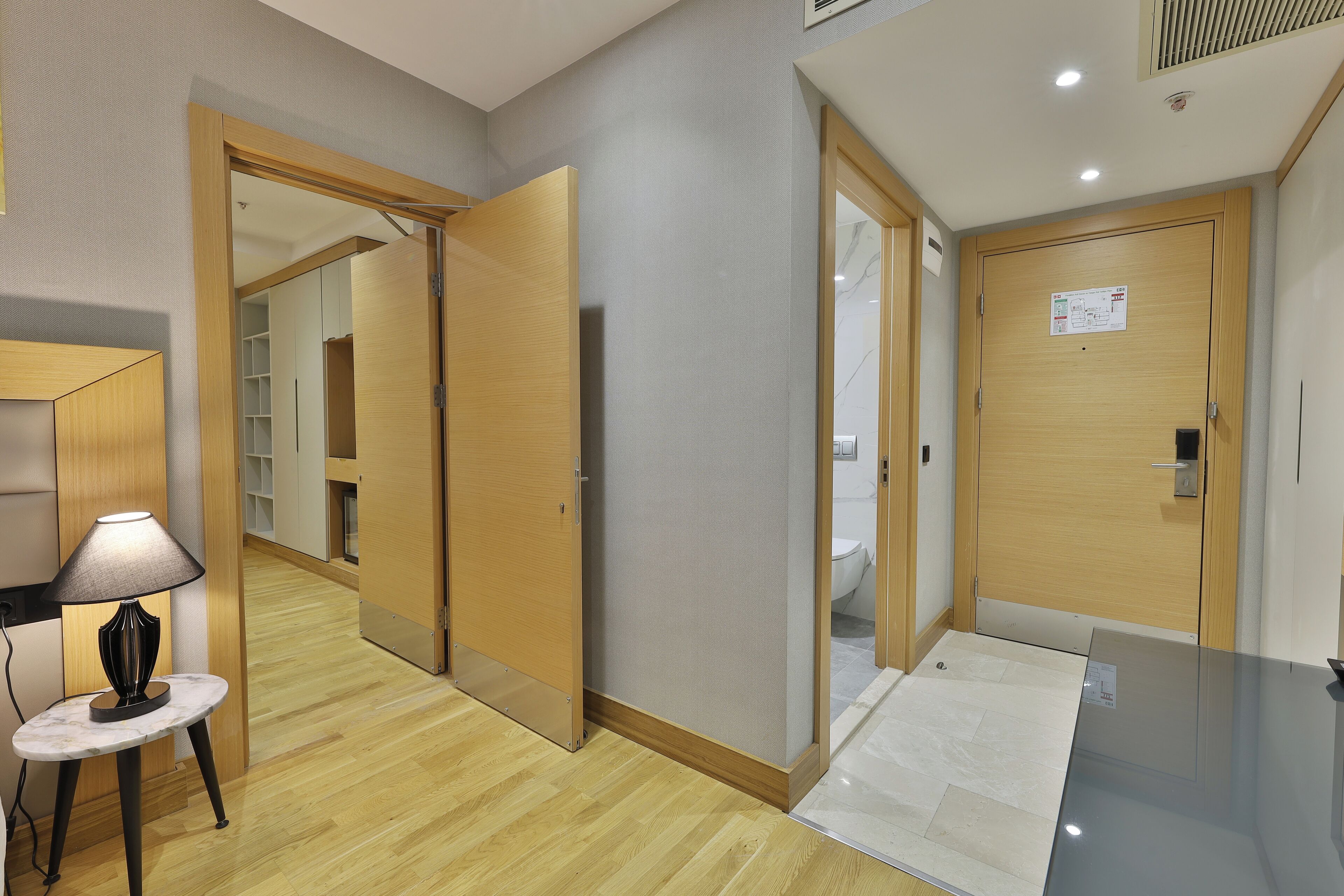 Un pasillo de hotel con puertas de madera, un espejo, una mesa con lámpara y suelo de baldosas.