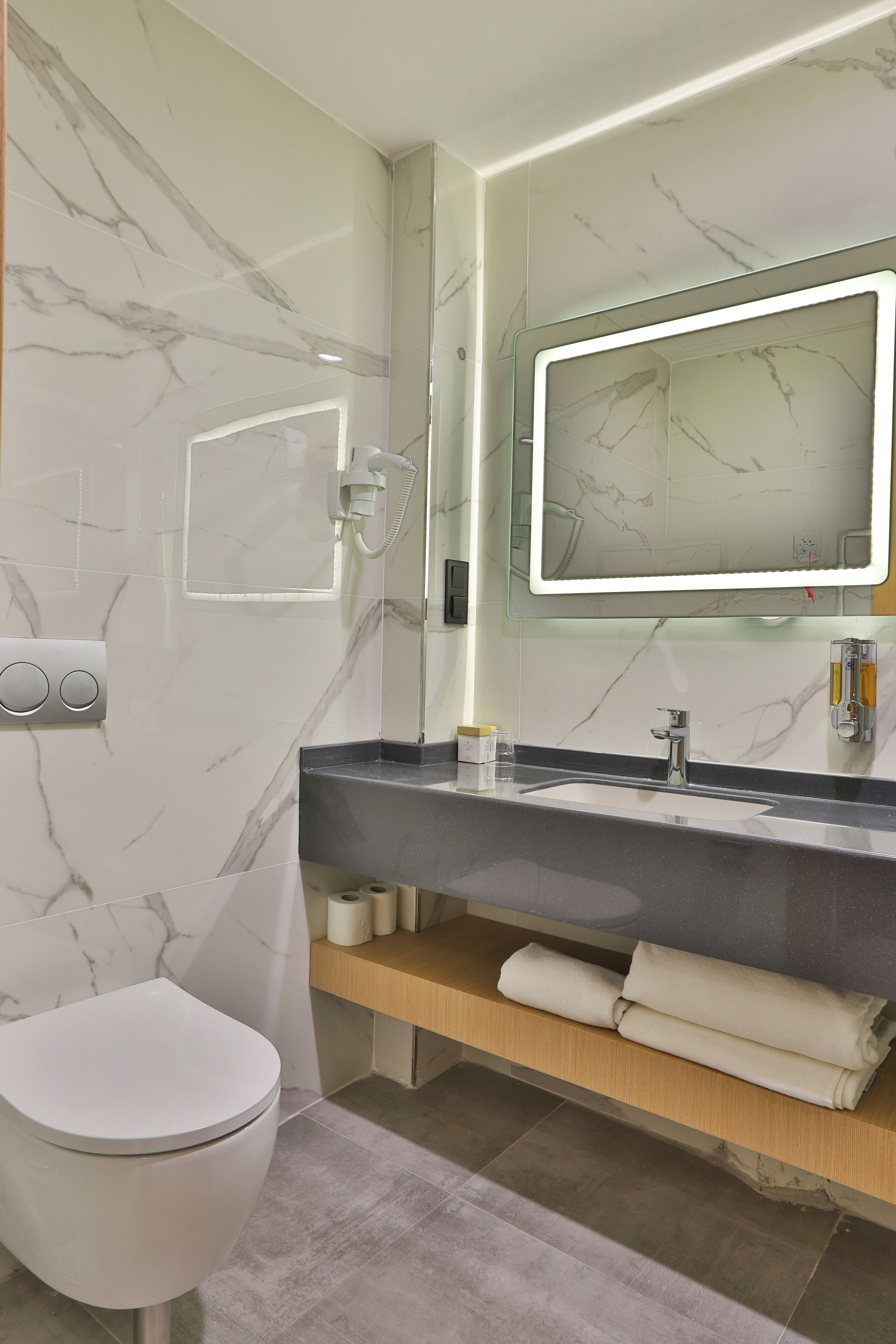 A modern bathroom with a marble wall, a large mirror, a sink with a towel holder, and a toilet.