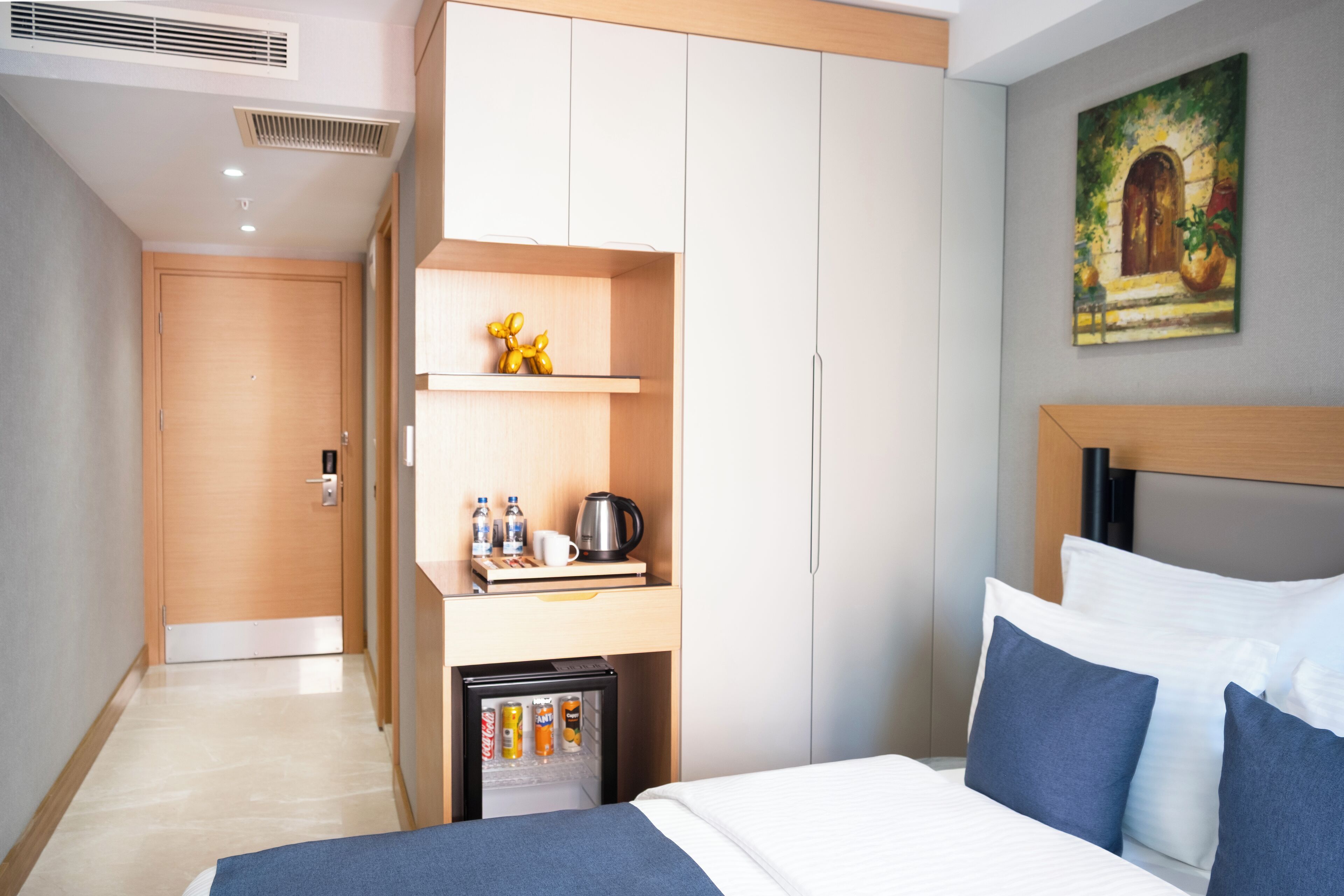 A hotel room with a bed, a wardrobe, a mini-fridge, and a painting on the wall.