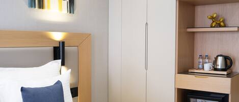 A hotel room with a bed, a nightstand, a mini-fridge, a shelf with bottles, and a painting on the wall.