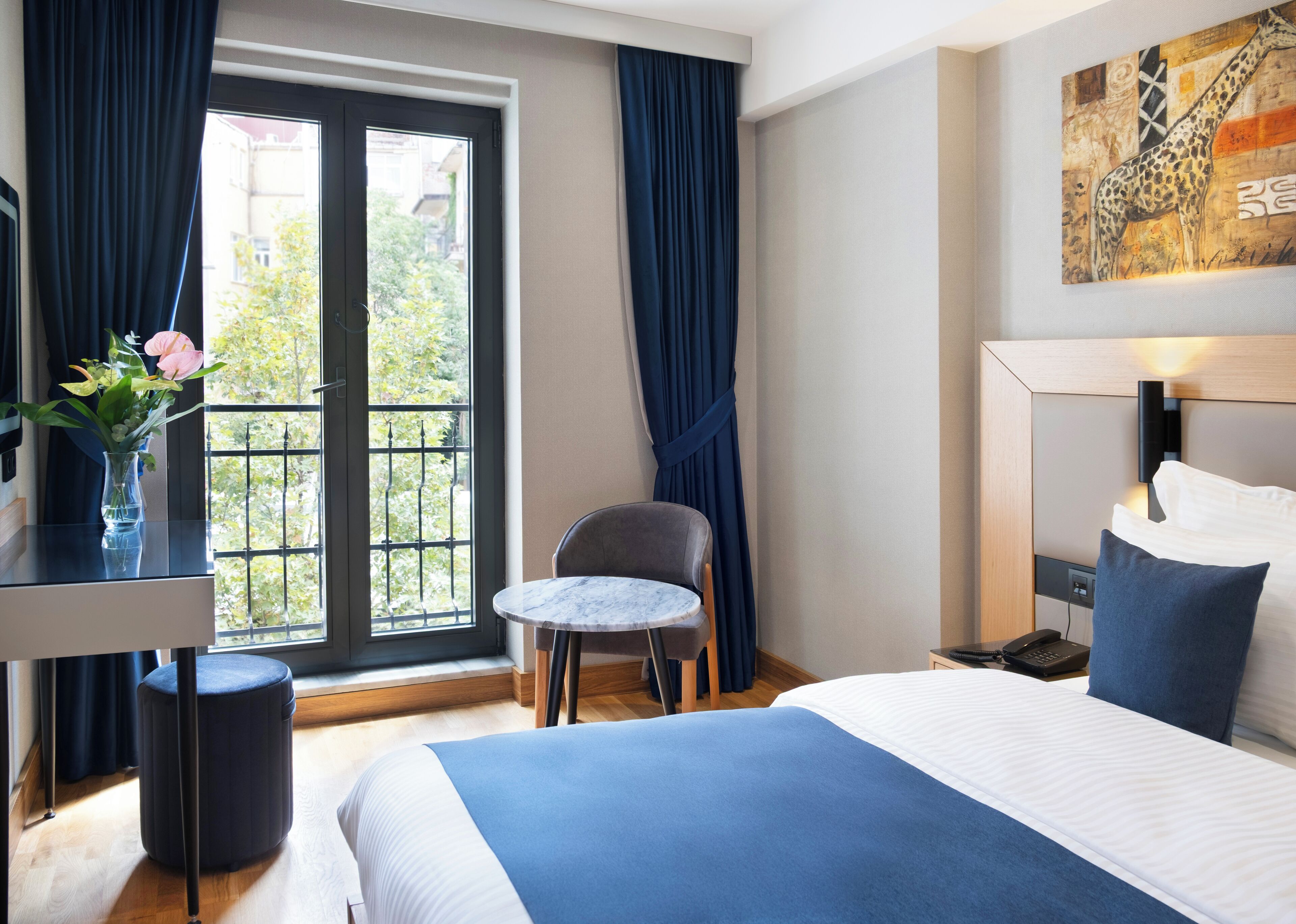 Habitación de hotel con cama, una mesita redonda, una silla, un escritorio con un florero y vistas al exterior a través de una ventana con cortinas.