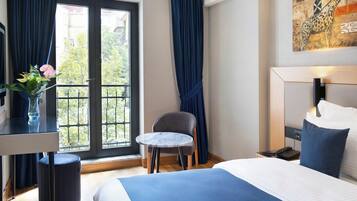 Une chambre d’hôtel avec un lit, une petite table ronde, une chaise, un bureau agrémenté d’un vase de fleurs et une vue sur l’extérieur par une fenêtre munie de rideaux.