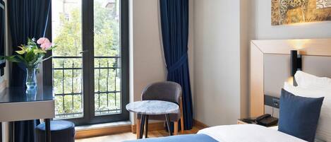 Habitación de hotel con cama, una mesita redonda, una silla, un escritorio con un florero y vistas al exterior a través de una ventana con cortinas.