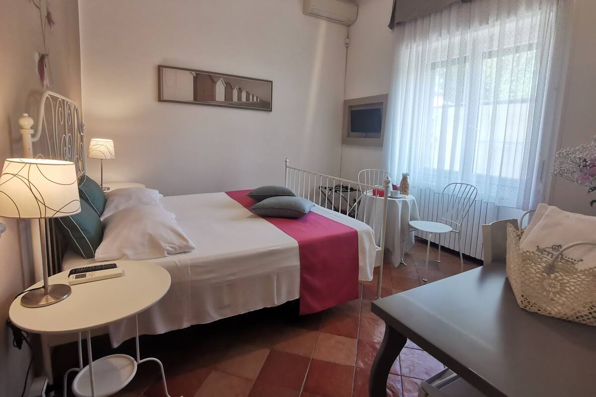 Een hotelkamer met een bed, nachtkastjes, een kleintje tafeltje, een stoel, een raam met gordijnen en een wandairconditioner.