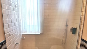 Un pequeño espacio de ducha cerrado con puerta de cristal, una ventana con cortinas translúcidas y un revestimiento de azulejos blancos.