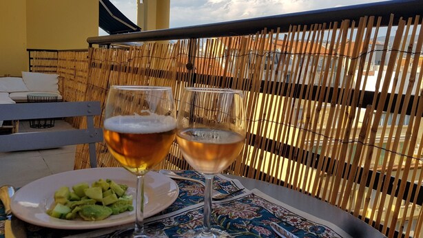 Une table avec deux verres de bière et une assiette de nourriture, placée devant une clôture en bambou, avec vue sur des bâtiments et un ciel nuageux.