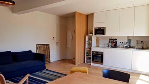 Un salon moderne avec un canapé bleu, une table basse en bois et un petit pouf jaune. On aperçoit, au fond, une cuisine avec des meubles blancs et un four à micro-ondes.