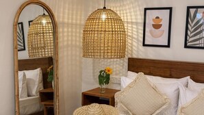 A bedroom with a wooden headboard, a large mirror, two woven pendant lights, a bedside table with a vase of flowers, and a bed with white linens and textured pillows. - Kickin' Surf Lodge (Weligama)