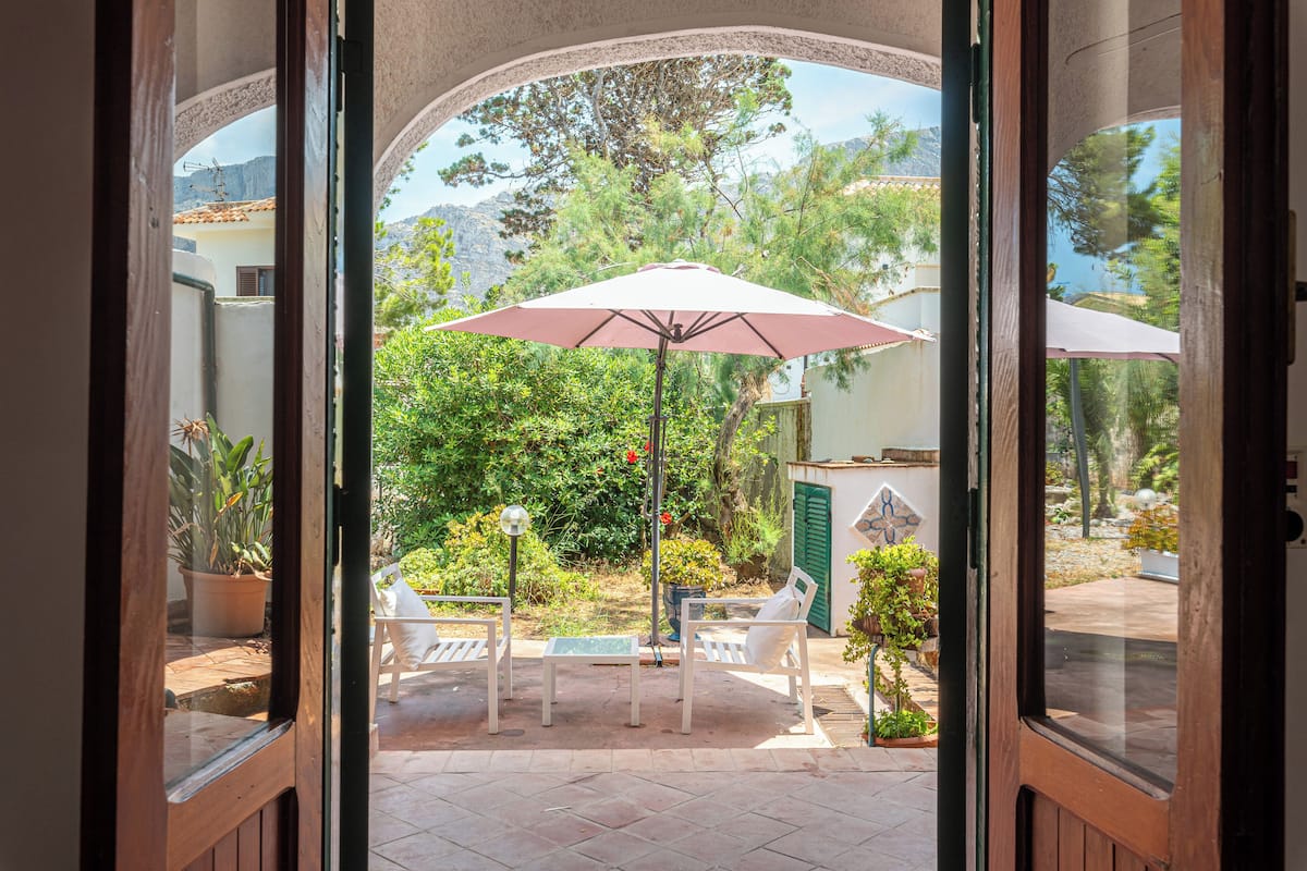 Une porte ouverte donne sur un patio aménagé avec une table et des chaises sous un parasol rose, entouré de plantes en pot et offrant une vue sur un paysage montagneux.