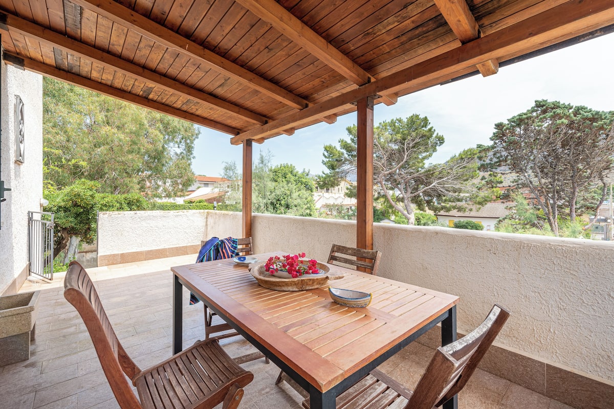 Une table en bois, des chaises et un vase de fleurs sont disposés sur un balcon d’où l’on peut admirer les arbres et les bâtiments.