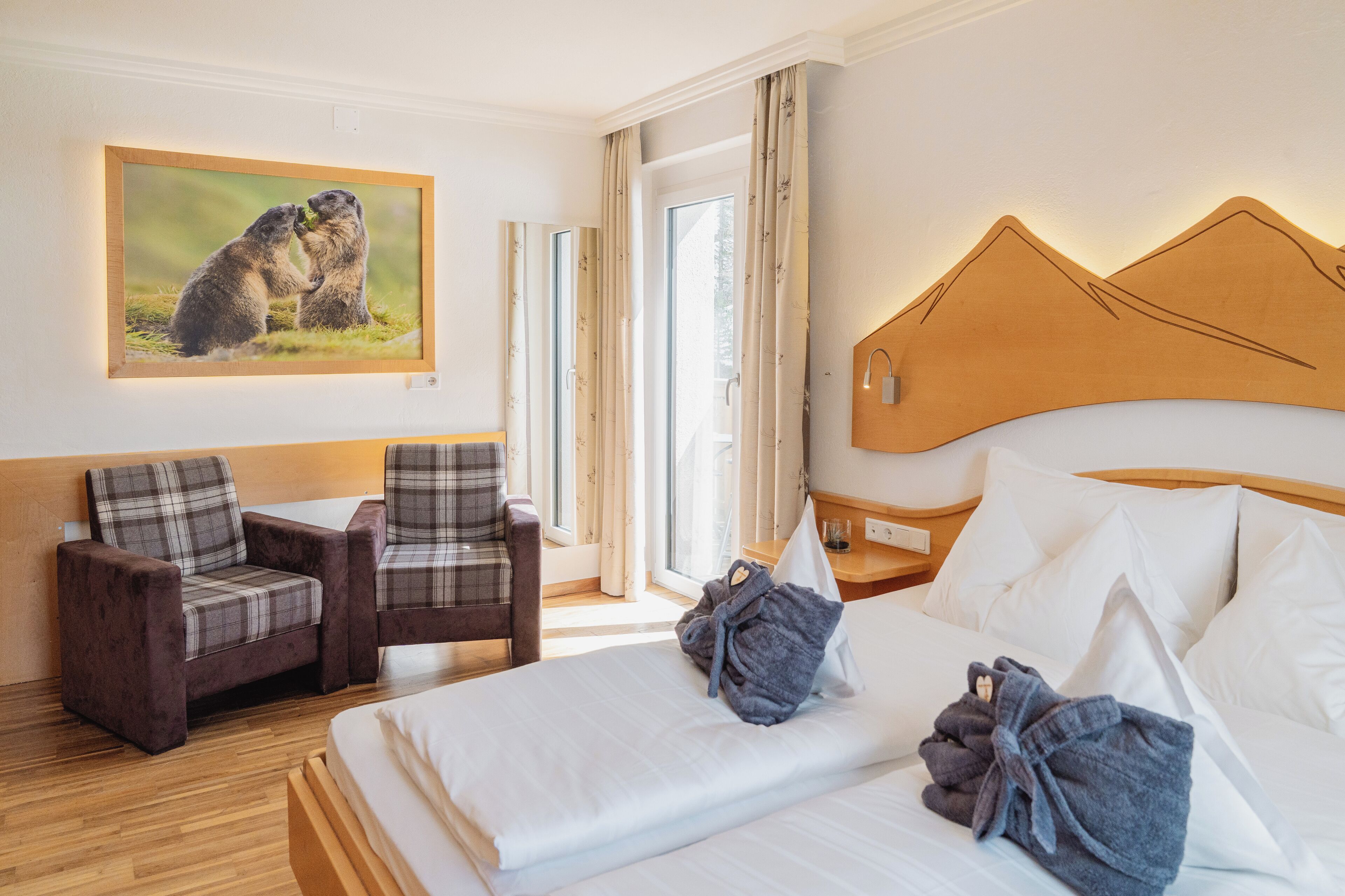 A hotel room with a bed, two armchairs, a painting of animals, and a window with curtains.