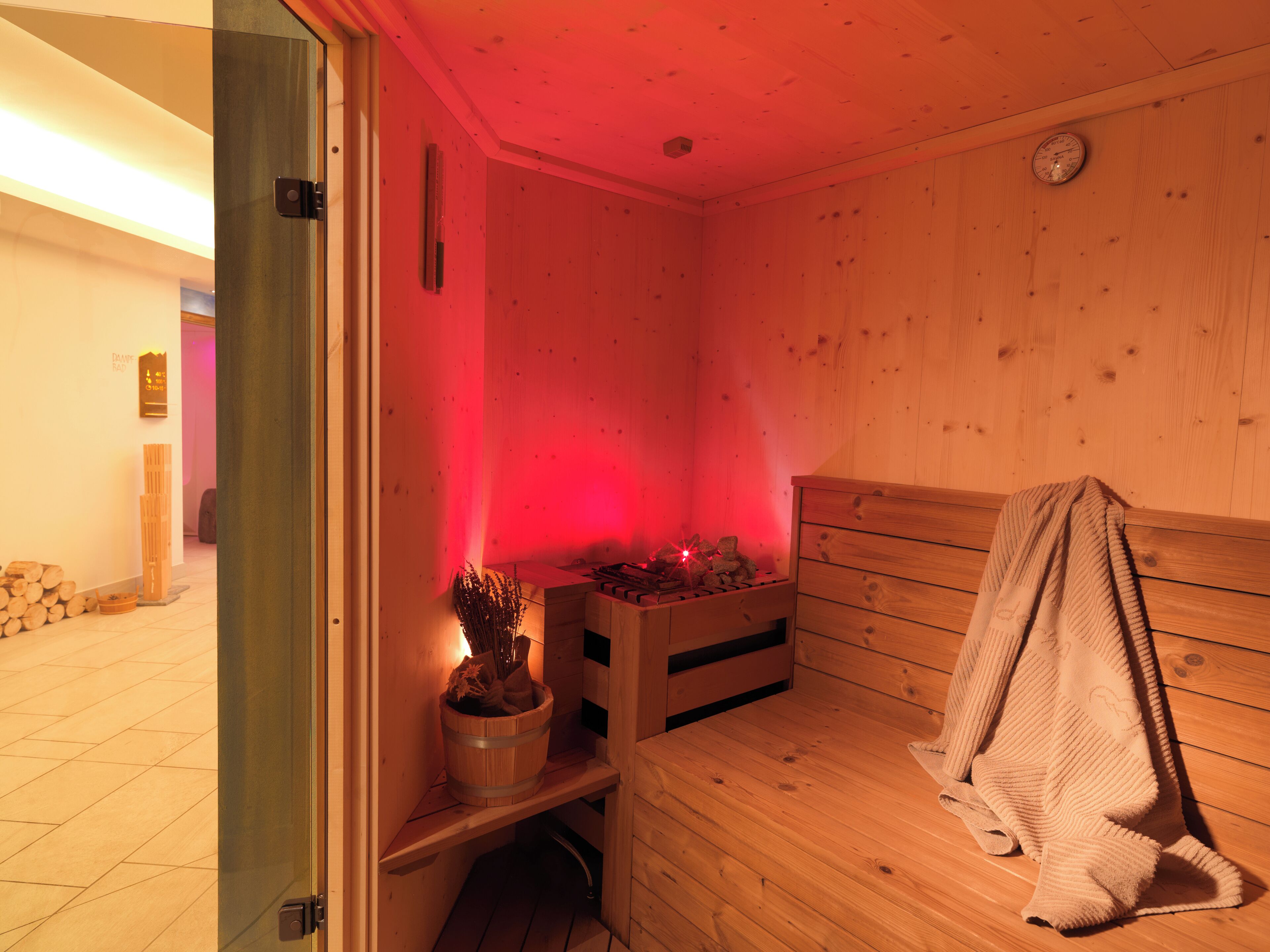 Un sauna con paredes de madera y un banco. Se observa un cubo de madera con una planta y una toalla colgada sobre el banco.