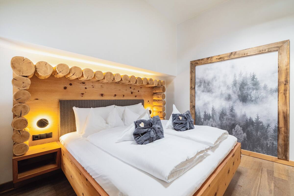 Una cama bien hecha con sábanas blancas y dos toallas, un cabecero de madera con una luz circular y un mural grande con motivos forestales.