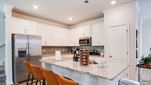A modern kitchen with white cabinets, a stainless steel refrigerator, a microwave, and a granite countertop. There are three orange bar stools and a stainless steel trash can.