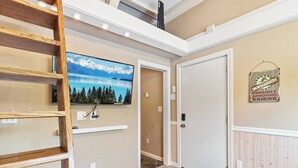 A hallway with a wooden ladder, a mounted TV displaying a mountain landscape, a door, and a wall sign reading "Snow Boarders Proudly Annoying Skiers".