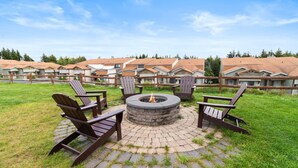 A circular fire pit surrounded by Adirondack chairs in a courtyard with a brick floor, set against a backdrop of residential buildings.