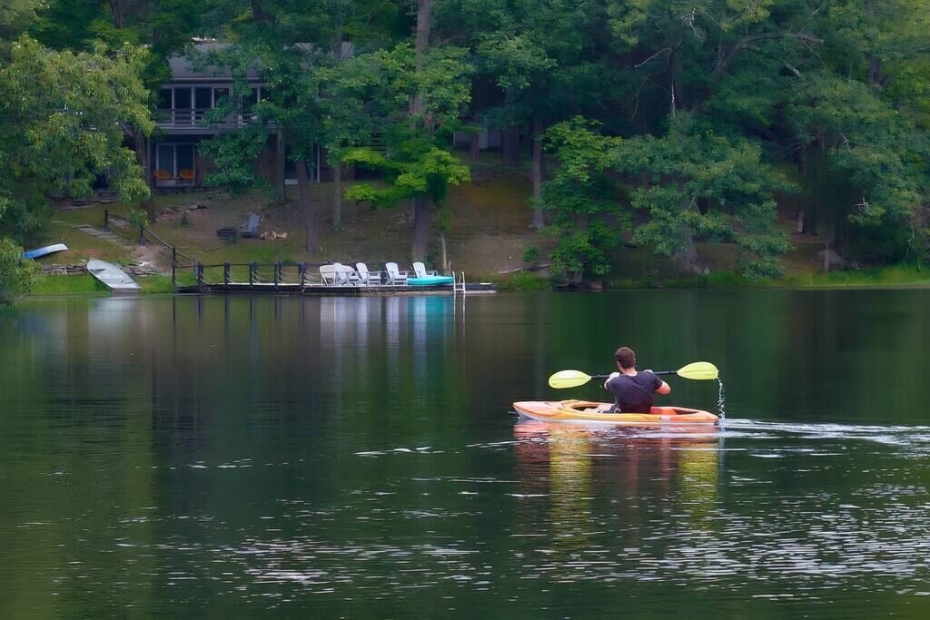 A person kayaking on a calm lake with a dock and lounge chairs in the background.