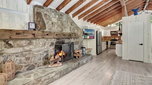 Un interior rústico con chimenea de piedra, vigas de madera, una zona de cocina y una puerta blanca.
