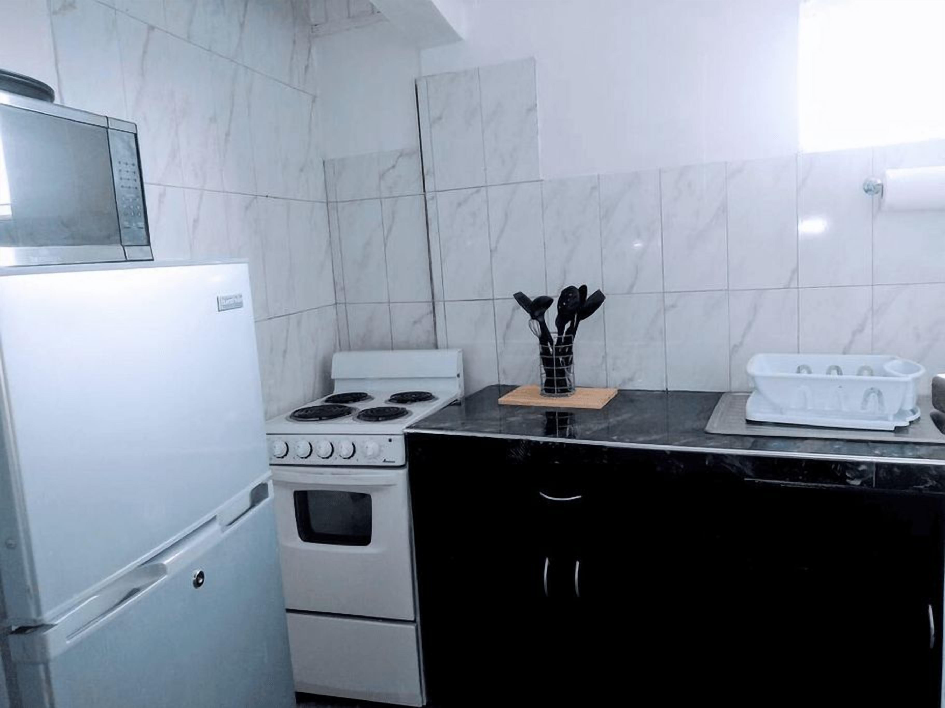 Cuina petita amb neveri, fogó, forn i microones. Hi ha un taulell negre amb un suport per ganivets i una safata blanca.