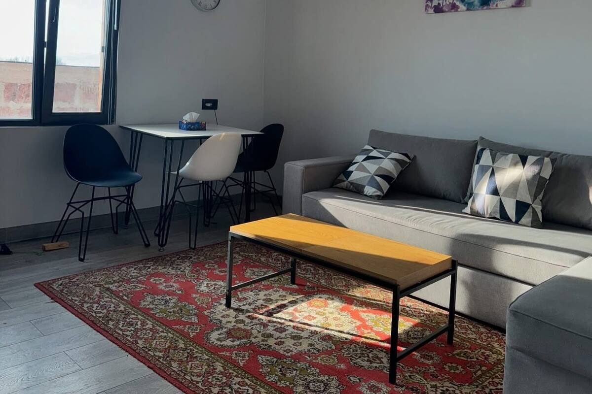 A modern living room with a grey sofa, a wooden coffee table, and a patterned rug. There is a dining area with a small table and chairs, and a large window providing natural light.