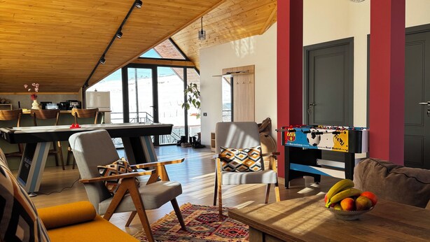 Amplia sala de estar con techo alto, vigas de madera y un ventanal grande. Hay un espacio para sentarse con un sofá y sillones, una mesa de centro con fruta, y una mesa de fútbol en el fondo.