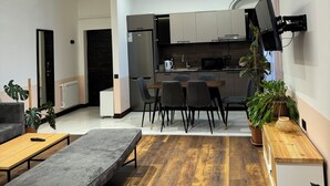 Una sala moderna con un sofá gris, una mesa de centro de madera y un banco gris. Hay una zona de cocina con una mesa de comedor y sillas, un microondas y un hervidor. El cuarto tiene suelo de madera y un ventanal con cortinas.