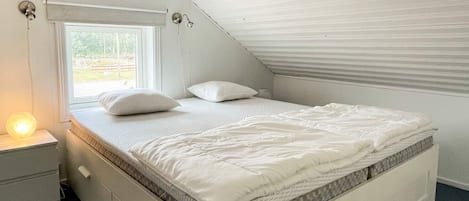 A bedroom with a sloped ceiling, a bed with white bedding, a nightstand with a lamp, and a window with a view of the outdoors.