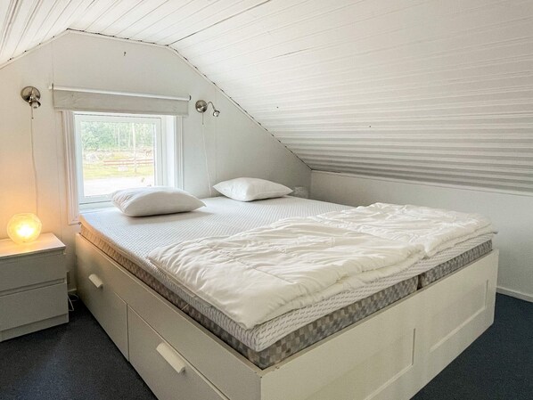 A bedroom with a sloped ceiling, a bed with white bedding, a nightstand with a lamp, and a window with a view of the outdoors.