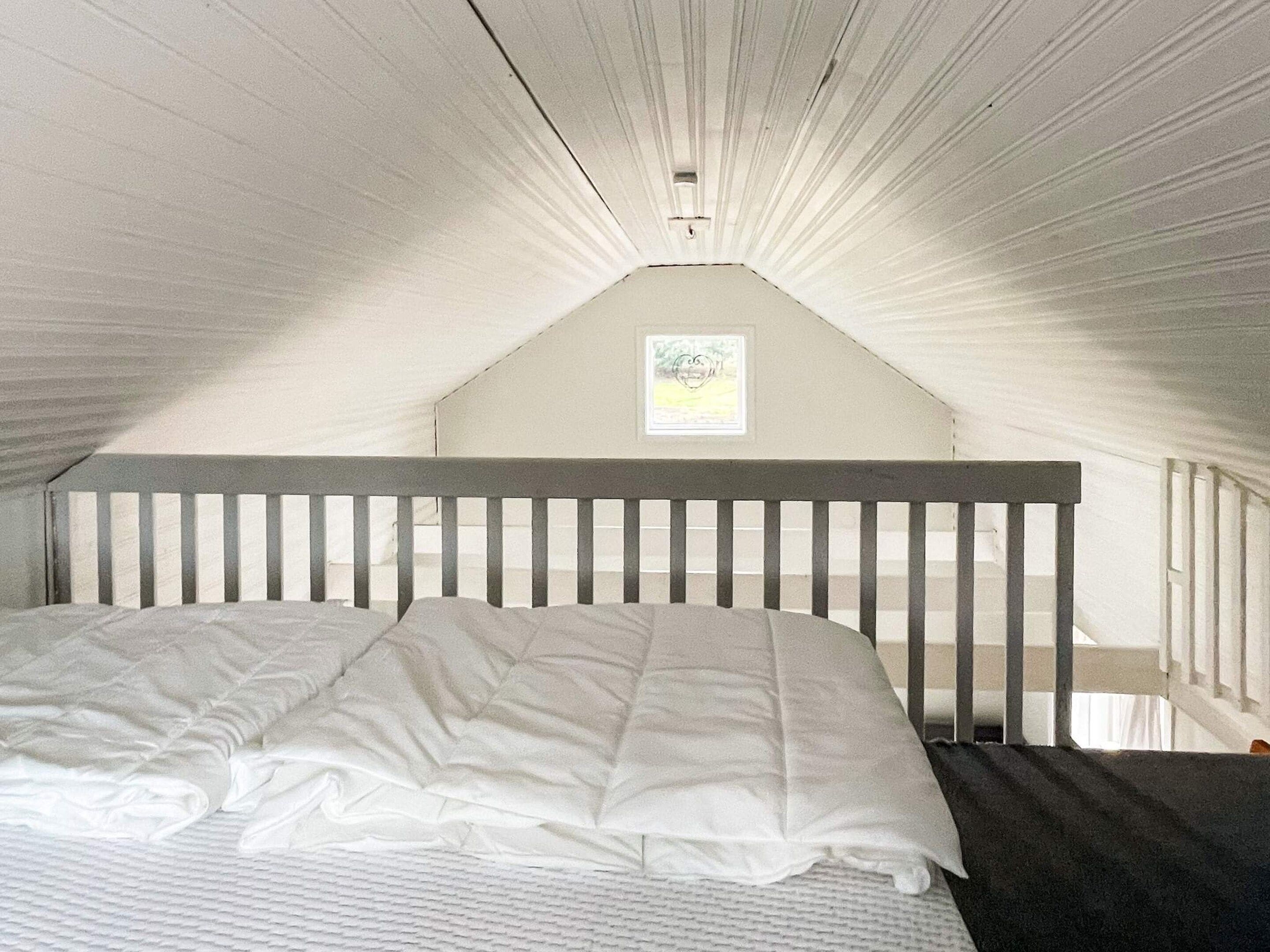 Une chambre avec un plafond en pente, un lit simple avec un matelas et des oreillers blancs, et une petite fenêtre ornée d’un tableau encadré.
