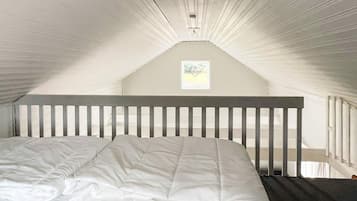 A bedroom with a sloped ceiling, a single bed with a white mattress and pillows, and a small window with a framed picture.