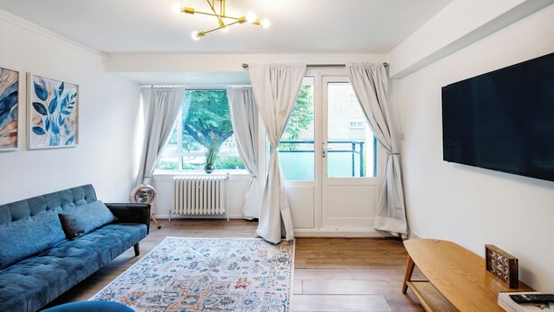 A modern living room with a blue sofa, a patterned rug, a wooden bench, a flat-screen TV, and a large window with curtains.