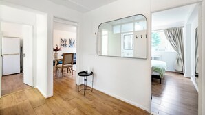 A room with a wooden floor, a bed with a green bedspread, a small table with a vase, and a large mirror on the wall.