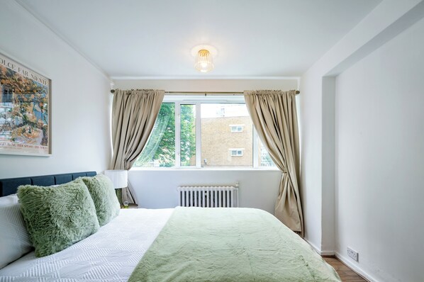A bedroom with a bed, a window with curtains, a radiator, and a wall-mounted light fixture. - Cozy 2 bedroom flat- Heart of Victoria (London)