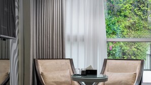 A modern room with two beige armchairs, a round glass table, and a view of greenery through large windows. - J Village Hotel - Triệu Việt Vương (Hanoi)