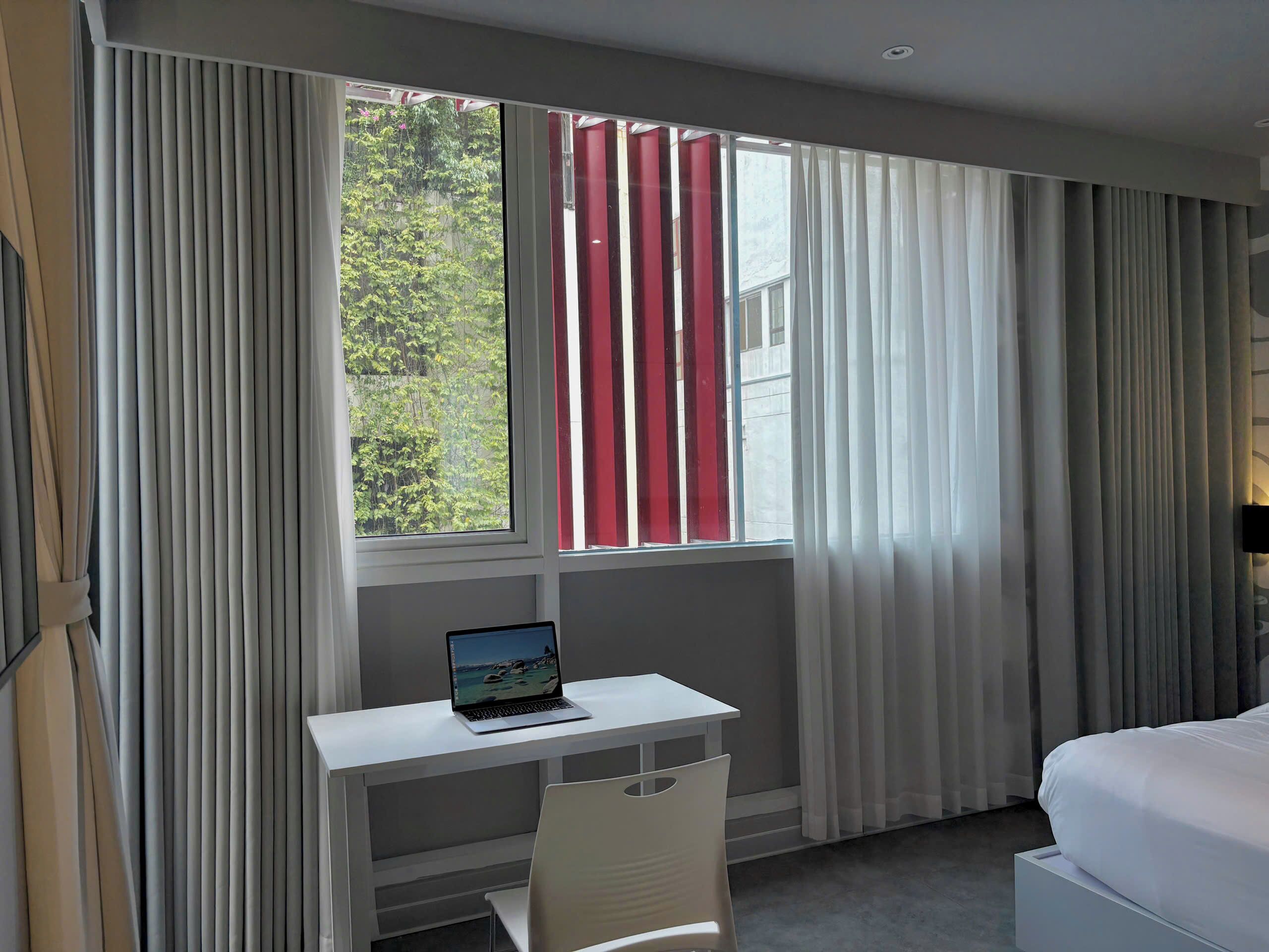 Ein Hotelzimmer mit einem Bett, einem Schreibtisch mit einem Laptop und einem Fenster mit Vorhängen.