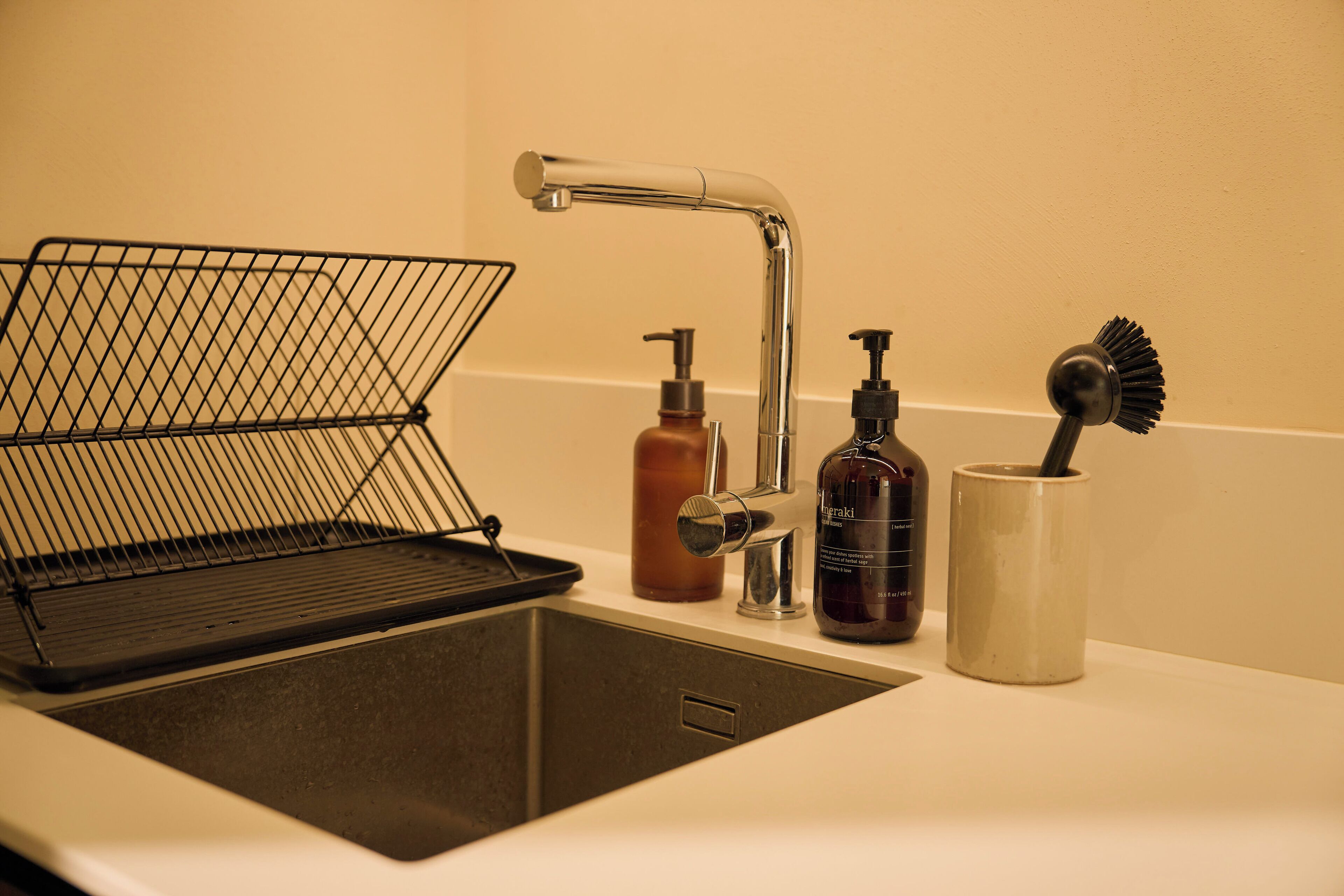 A kitchen sink with a wire basket, a soap dispenser, a bottle of soap, and a brush in a cup.