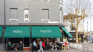 A café with outdoor seating, a green awning, and a sign indicating it serves artisan baked goods and British charcuterie.