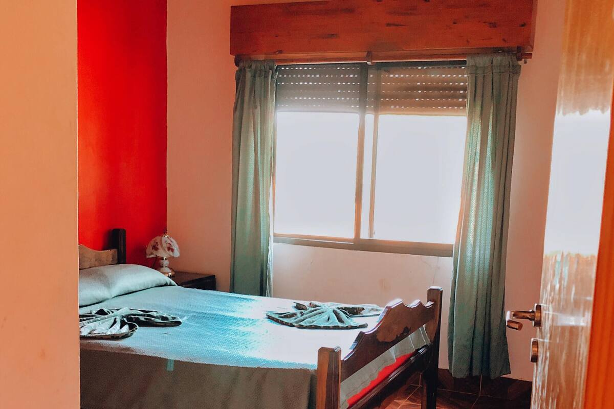 A bedroom with a bed, a wooden ceiling fan, a window with curtains, and a tiled floor.