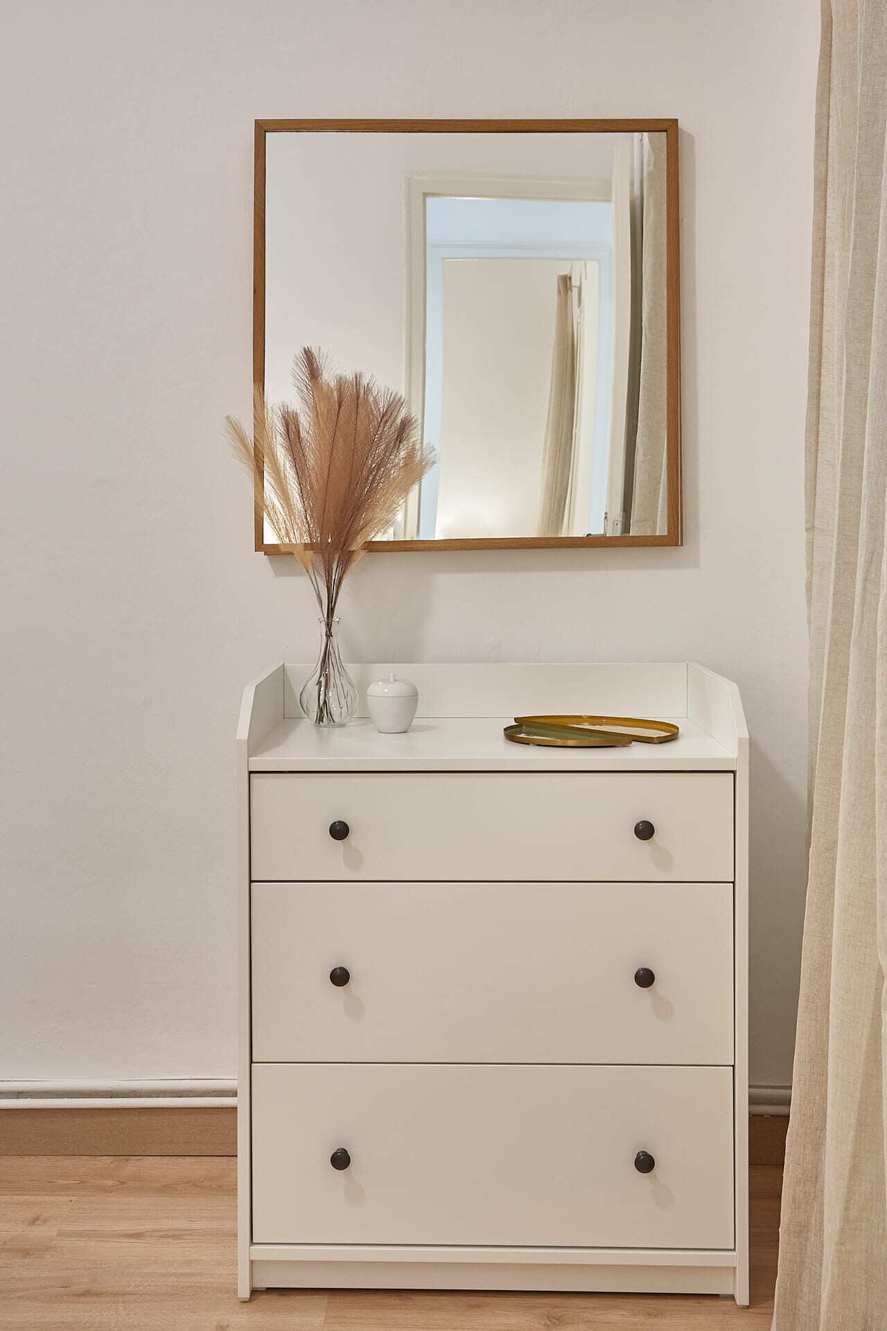 Een witte dressoir met drie lades, een spiegel erboven en een vaas met gedroogde planten erop.