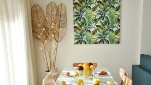 Een eethoek met een witte tafel, houten stoelen, een schaal met fruit en een wanddecoratie met een tropisch bladpatroon.