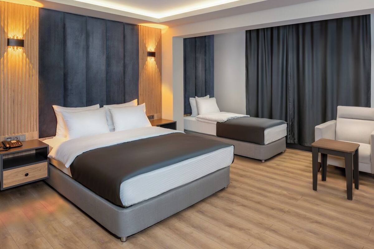 A modern hotel room with a large bed, a sofa, a chair, and a small table. There are two nightstands with lamps and a phone.